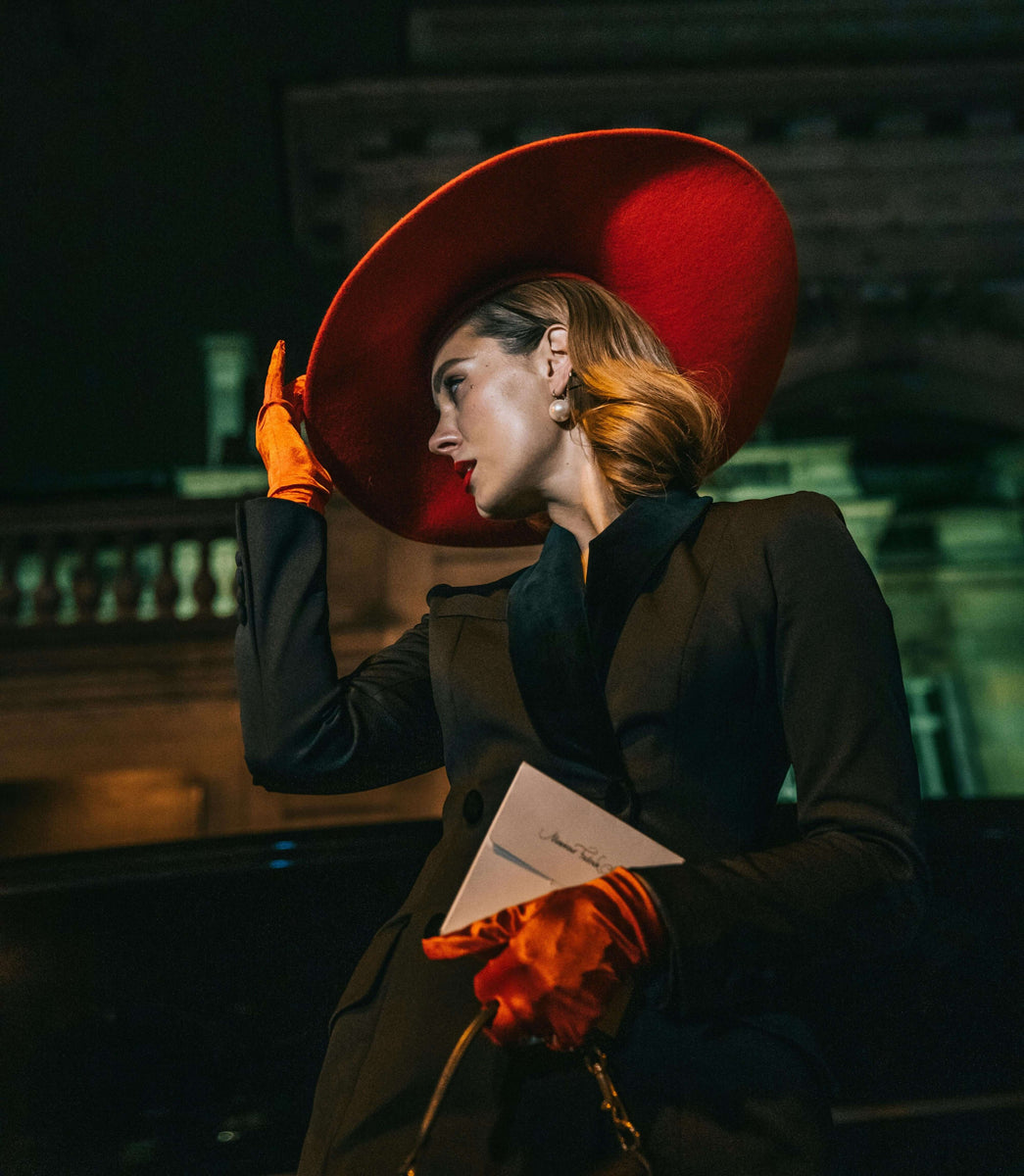 Elegant woman in black suit and red hat holding paper, posed at night against a historic building backdrop.